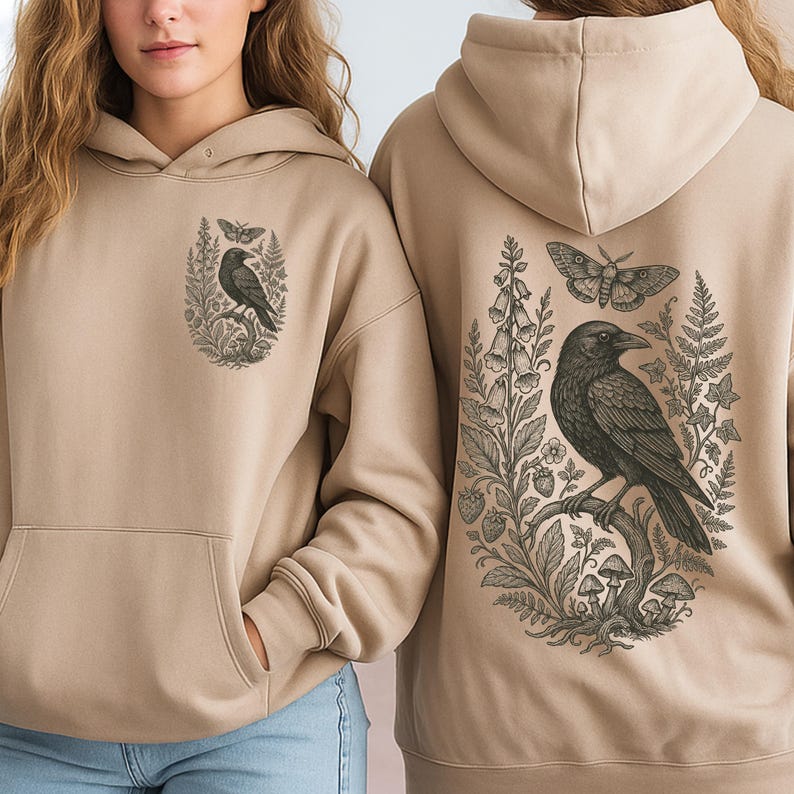 May include: A beige hooded sweatshirt featuring a black ink-style illustration of a raven perched on a branch, surrounded by ferns, mushrooms, and a moth. The front chest displays a smaller version of the same raven and moth graphic design.
