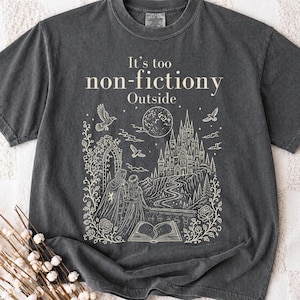 May include: A charcoal grey short-sleeve t-shirt featuring a cream-coloured line art graphic of a castle, a moon, birds, roses, an open book, and two figures. The text "It's too non-fictiony Outside" is printed in a serif font above the illustration.