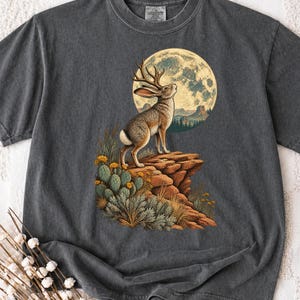 Pode incluir: Uma t-shirt cinzento-carvão com uma estampa gráfica de um jackalope em cima de um penhasco rochoso no deserto. A cena inclui uma lua cheia grande ao fundo, cactos com flores amarelas e ervas secas em tons terrosos.