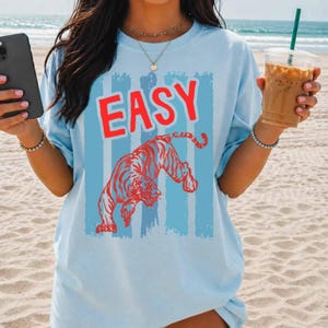 May include: A light blue short-sleeve t-shirt featuring a graphic print of a red tiger and the word "EASY" in red capital letters. The design is set against a background of vertical light blue stripes. The shirt is worn with a gold necklace.