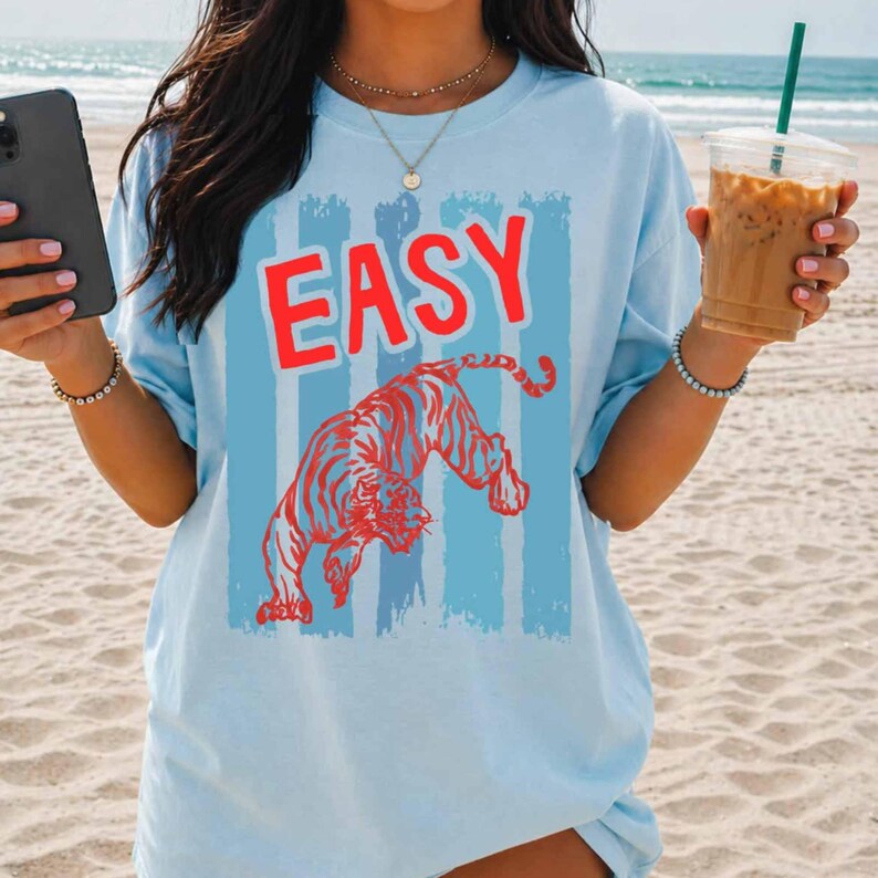 May include: A light blue short-sleeve t-shirt featuring a graphic print of a red tiger and the word "EASY" in red capital letters. The design is set against a background of vertical light blue stripes. The shirt is worn with a gold necklace.