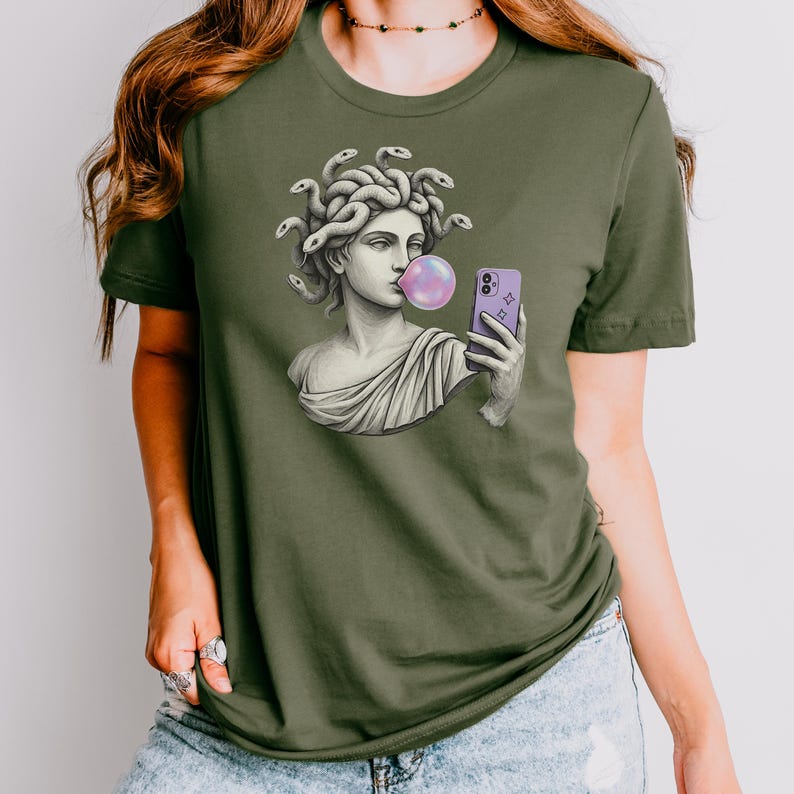 May include: Olive green t-shirt with a graphic of a Medusa bust blowing a bubblegum bubble while holding a purple smartphone. The Medusa bust is in a classical style, with snakes for hair. The bubble is pink.