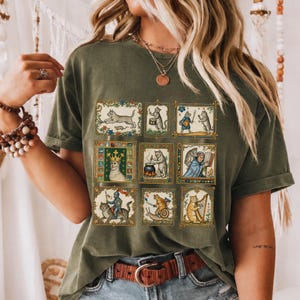 Op de afbeelding: Een olijfgroen t-shirt met een raster van negen rechthoekige illustraties van antropomorfe katten in middeleeuwse sc&egrave;nes, waaronder een kat met een kroon, een kat die harp speelt en een kat op een slak, allemaal omlijst door decoratieve gouden randen.