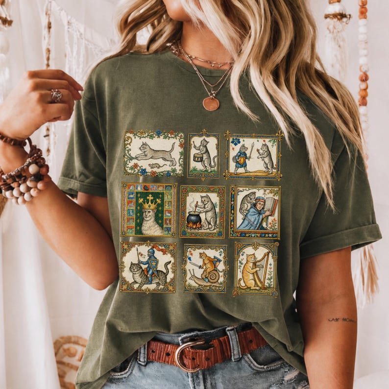 Op de afbeelding: Een olijfgroen t-shirt met een raster van negen rechthoekige illustraties van antropomorfe katten in middeleeuwse sc&egrave;nes, waaronder een kat met een kroon, een kat die harp speelt en een kat op een slak, allemaal omlijst door decoratieve gouden randen.