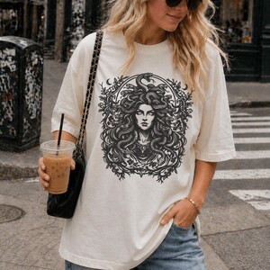 Op de afbeelding: Cr&egrave;mekleurig T-shirt met een zwart-witte illustratie van Medusa, met slangen als haar, omringd door een bloemenkrans. Het shirt heeft korte mouwen en een relaxte pasvorm.