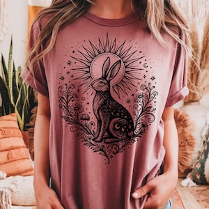 Peut inclure: Un t-shirt rose poudré avec une illustration noire détaillée d'un lapin, entouré de fleurs, d'étoiles et d'un motif de soleil. Le graphisme est centré sur le t-shirt, créant une esthétique bohème ou inspirée de la nature. Le t-shirt a des manches courtes et une coupe décontractée.