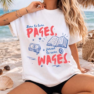 May include: White t-shirt with the text "Born to turn PAGES, forced to earn WAGES." The design includes illustrations of a stack of money, an open book, and a coin. The text and illustrations are in red and blue.