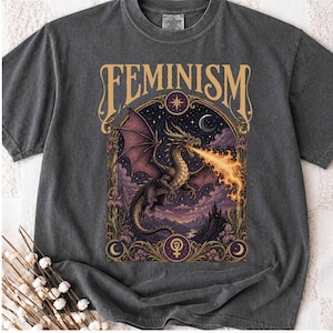 Op de afbeelding: Een antracietgrijs t-shirt met ronde hals en een grafische print met de tekst FEMINISM bovenaan. Het ontwerp toont een vuurspuwende draak binnen een sierlijk boogframe, omringd door paarse wolken, een maansikkel en een ster.