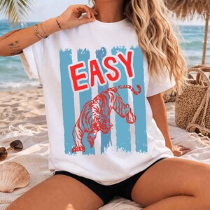 May include: A white short-sleeve t-shirt featuring a graphic print with the word EASY in bold red letters above a stylized red tiger illustration. The design is set against a background of vertical light blue distressed stripes on the front.