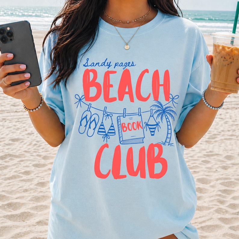 May include: Light blue t-shirt with the words "Sandy pages BEACH CLUB" in red. The design includes illustrations of flip-flops, a book, a palm tree, and a bikini. The shirt is made of a soft material.
