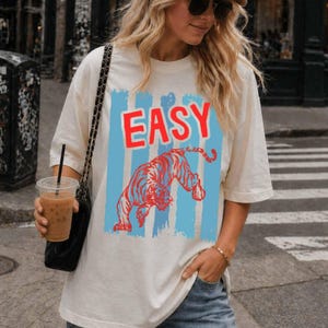May include: An off-white short-sleeve t-shirt featuring a graphic print with the word EASY in bold red letters above a red tiger illustration, all set against a background of vertical light blue brushstroke stripes on the front of the garment.