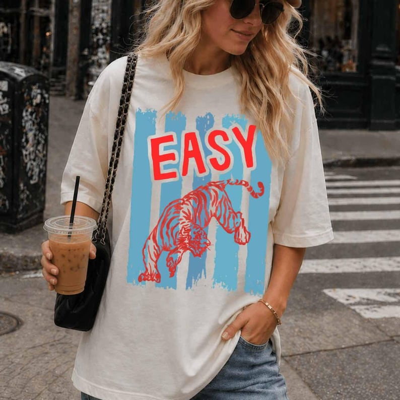 May include: An off-white short-sleeve t-shirt featuring a graphic print with the word EASY in bold red letters above a red tiger illustration, all set against a background of vertical light blue brushstroke stripes on the front of the garment.