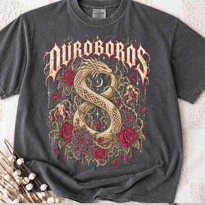 May include: A charcoal grey short-sleeve t-shirt featuring a graphic print of a golden serpent coiled into an infinity shape. The design includes red roses, thorny vines, and the text DUROBOROS in a stylised, weathered font across the top.