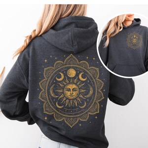 Puede incluir: Sudadera con capucha gris oscuro con un dise&ntilde;o de mandala dorado de sol y luna en la espalda. El dise&ntilde;o incluye un sol sonriente, lunas crecientes y estrellas. Una versi&oacute;n m&aacute;s peque&ntilde;a del dise&ntilde;o est&aacute; en la parte delantera.