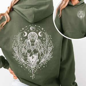 Puede incluir: Sudadera con capucha verde oliva con un diseño blanco en la espalda que representa a una mujer sosteniendo una llave, rodeada de elementos celestiales. El diseño incluye lunas, estrellas y detalles florales. Un diseño más pequeño está en el frente.