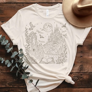 May include: A cream-coloured short-sleeve t-shirt featuring a dark brown line art illustration of a castle, a full moon, birds, and two figures standing before an open book, all framed by floral vines and a stone archway on the front.