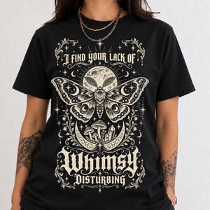 Op de afbeelding: Een zwart t-shirt met een crèmekleurige afbeelding van een grote mot boven een halve maan, paddenstoelen en kristallen. De tekst luidt I FIND YOUR LACK OF WHIMSY DISTURBING, omringd door decoratieve florale en hemelse ster-elementen.