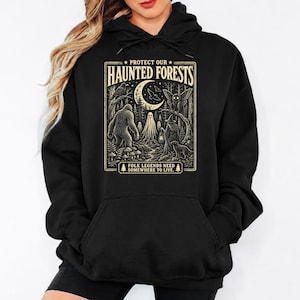 Puede incluir: Una sudadera con capucha negra con un estampado gráfico de color crema. El diseño muestra una escena forestal con una luna creciente, una criatura grande, un fantasma, una polilla y una figura con una linterna. El texto dice: PROTECT OUR HAUNTED FORESTS FOLK LEGENDS NEED SOMEWHERE TO LIVE.