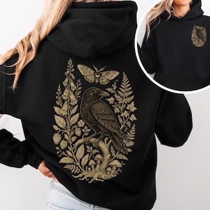 May include: A black hooded sweatshirt featuring a large gold-toned graphic on the back depicting a raven perched on a branch surrounded by mushrooms, ferns, foxgloves, strawberries, and a moth. A smaller version of the raven graphic is on the front.