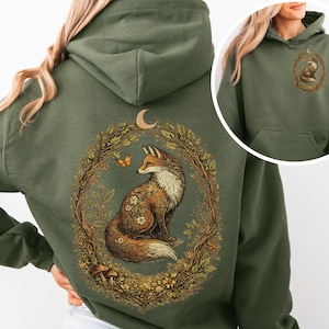 Puede incluir: Sudadera con capucha verde oliva con una ilustración detallada de un zorro dentro de una corona floral, una luna creciente y una mariposa. El zorro tiene patrones florales en su pelaje. Una versión más pequeña del diseño está en la parte delantera. La sudadera tiene capucha y bolsillo delantero.