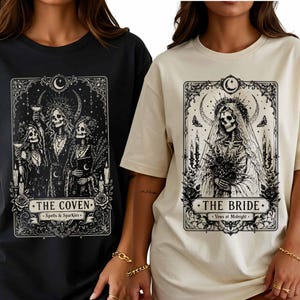 May include: A black t-shirt featuring a graphic of three skeletons labelled THE COVEN Spells & Sparkles and a cream t-shirt featuring a graphic of a skeleton bride labelled THE BRIDE Vows at Midnight, both in a tarot card style frame.