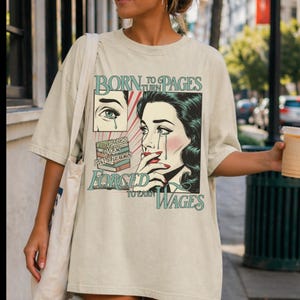 May include: Beige oversized t-shirt with a comic book style graphic. The graphic shows a crying woman and the text "BORN TO TURN PAGES, FORCED TO EARN WAGES". The design includes teal and red accents.