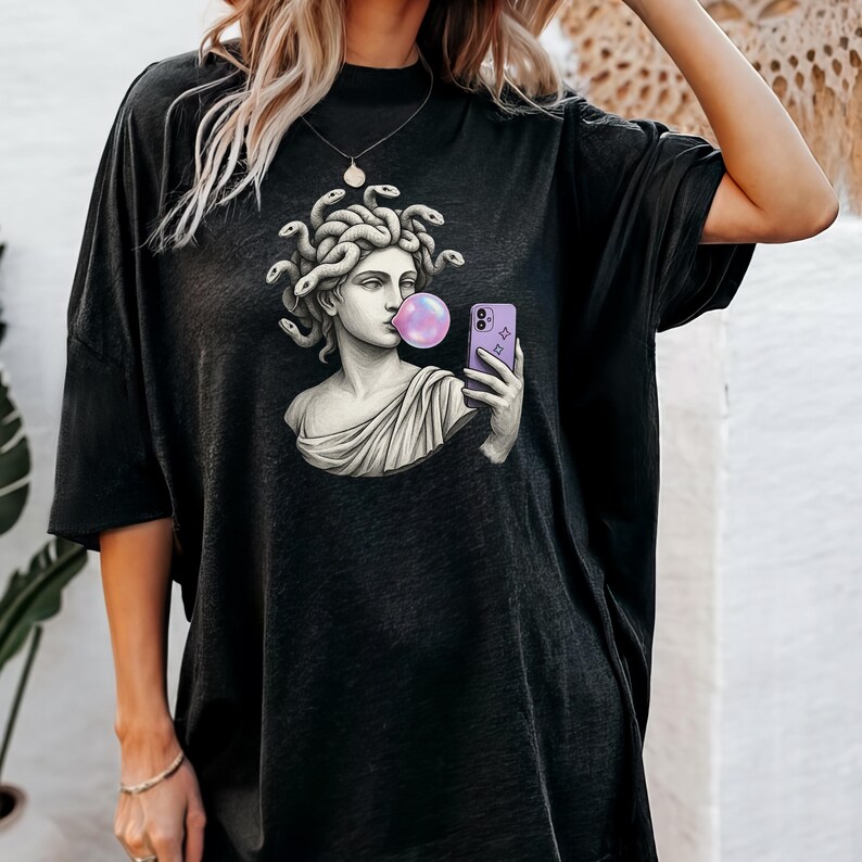 May include: Black oversized t-shirt with a graphic of a Medusa bust blowing a bubblegum bubble while taking a selfie with a purple phone. The Medusa bust is in a classical style with snakes for hair.