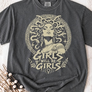May include: A charcoal grey short-sleeve t-shirt featuring a cream-coloured graphic of Medusa surrounded by snakes within a circular border. Below the image, the text reads GIRLS WILL BE GIRLS, accompanied by a female gender symbol and laurel leaves.