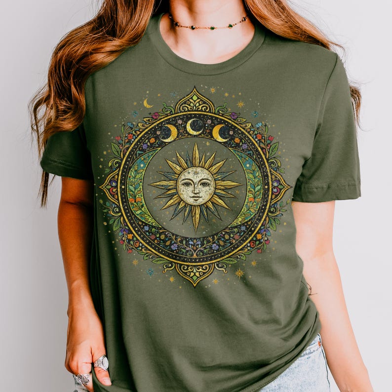 May include: Olive green t-shirt featuring a detailed sun and moon design. The central sun has a face, surrounded by crescent moons, floral elements, and a decorative border. The design is in shades of gold, green, and blue.