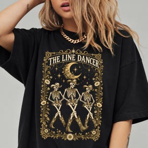 May include: A black t-shirt featuring a gold graphic print of three skeletons wearing cowboy hats and boots, dancing in a line. The design is framed by floral borders and a crescent moon, with the text THE LINE DANCER printed at the top.