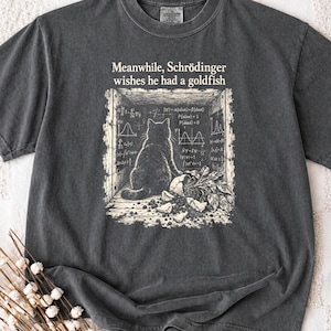 Op de afbeelding: Een antracietgrijs t-shirt met korte mouwen en een crèmekleurige afbeelding van een kat die in een doos zit met wiskundige vergelijkingen en de tekst "Meanwhile, Schrödinger wishes he had a goldfish" boven de illustratie gedrukt.
