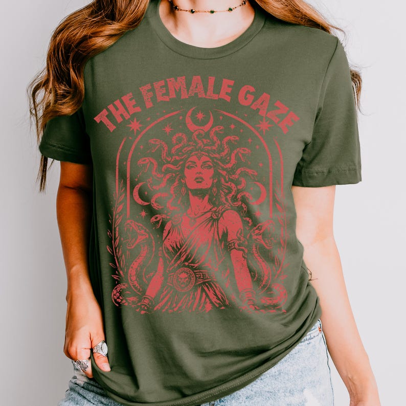 Pu&ograve; includere: T-shirt verde oliva con grafica rossa di Medusa e la scritta "THE FEMALE GAZE". La grafica presenta Medusa con serpenti al posto dei capelli, circondata da serpenti e lune crescenti. Il design &egrave; in stile vintage.
