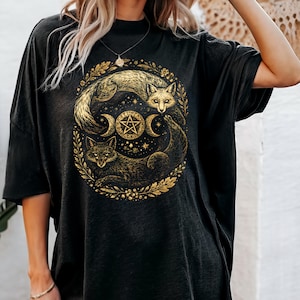 May include: Black t-shirt featuring a gold illustration of two foxes, crescent moons, and a pentagram within a circular floral design. The shirt has a relaxed fit and short sleeves.