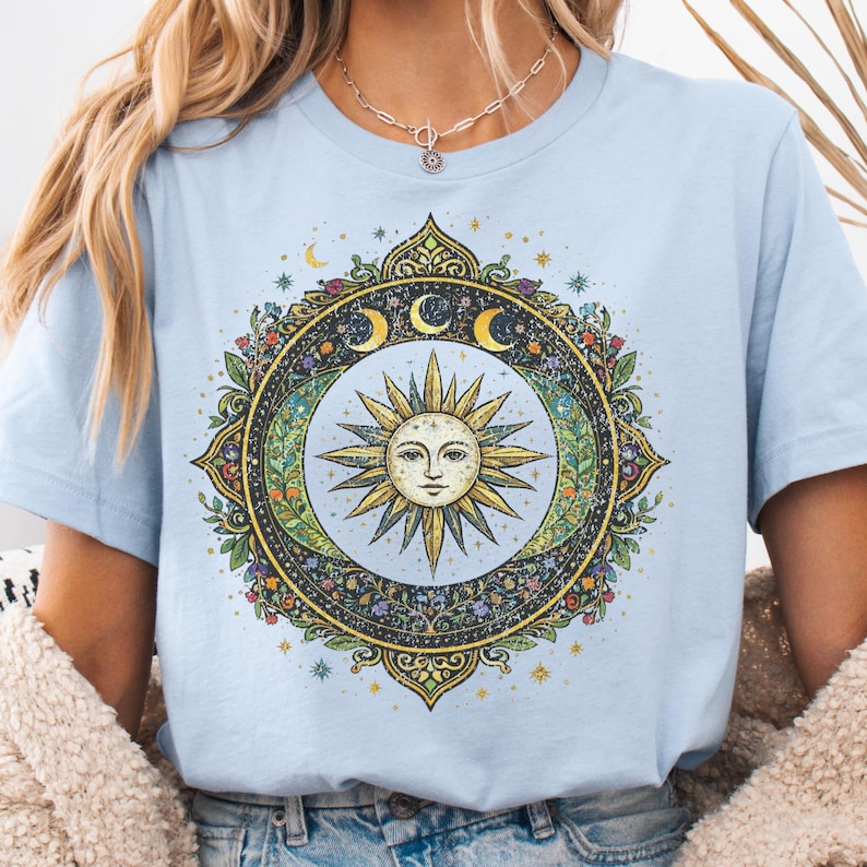 May include: Light blue t-shirt featuring a detailed circular design with a sun face at the center, surrounded by moons, stars, and floral elements. The design is in shades of gold, green, and blue.