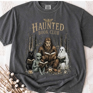 Op de afbeelding: Een antracietgrijs t-shirt met een grafische print met de tekst "HAUNTED BOOK CLUB" boven een illustratie van een Bigfoot, een alien, een zwarte kat en een geest die in een bos zitten, boeken lezen en uit kleine kopjes drinken.