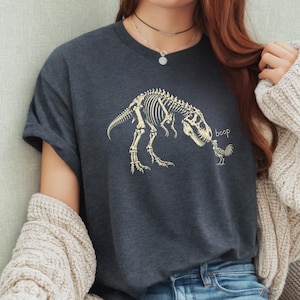 May include: A dark charcoal gray short-sleeve t-shirt featuring a light beige graphic print of a Tyrannosaurus Rex skeleton leaning down to touch a small chicken skeleton with its snout, accompanied by the lowercase text "boop" in beige.