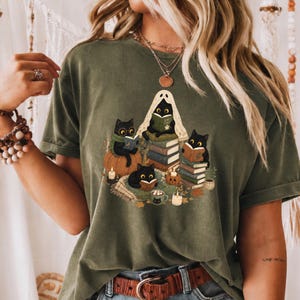 Pode incluir: Uma t-shirt de manga curta verde-azeitona com uma ilustração gráfica de quatro gatos pretos a ler livros. Um gato usa um lençol de fantasma, outro usa óculos e um cachecol verde, rodeados por pilhas de livros, abóboras e velas.