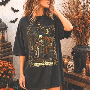 Pode incluir: Uma t-shirt preta com uma ilustração gráfica de um esqueleto sentado numa poltrona a ler um livro, acompanhado por um gato, pilhas de livros e uma lua crescente. O texto THE INTROVERT está impresso numa faixa decorativa na base.