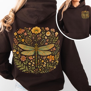 Può includere: Felpa con cappuccio marrone scuro con un design di libellula e fiori colorati sul retro. La libellula ha ali verdi e arancioni. Un design più piccolo è sulla tasca anteriore.