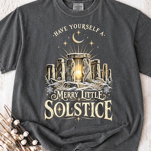 Może przedstawiać: Ciemnoszary t-shirt z grafiką przedstawiającą Stonehenge, świecące słońce między kamieniami, półksiężyc i gwiazdy. Napis brzmi, Have yourself a merry little solstice, wykonany ozdobną czcionką szeryfową.