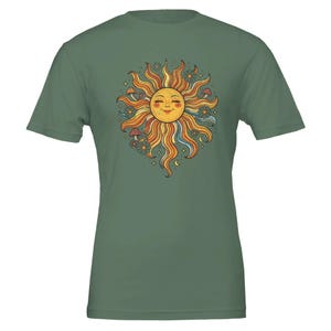 以下が含まれることがあります： セージグリーンの半袖Tシャツ。中央には、波打つ光線を持つ笑顔の太陽のグラフィックが描かれ、その周囲にはオレンジ、黄色、青、赤の色合いの小さなキノコ、花、星が配置されています。デザインは円形で中央に配置されています。