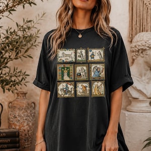 Op de afbeelding: Een zwart t-shirt met korte mouwen voorzien van een raster van negen rechthoekige illustraties met antropomorfe katten in middeleeuwse sc&egrave;nes, waaronder een kat als ridder, een kat die harp speelt en een kat bij een kookpot.