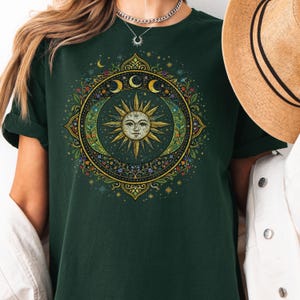 May include: Dark green t-shirt featuring a celestial design with a smiling sun, crescent moons, and floral accents. The design is circular and detailed, with gold and colorful elements. The shirt has short sleeves and a relaxed fit.