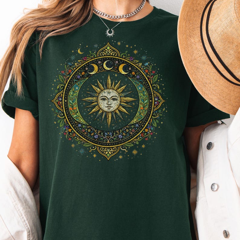 May include: Dark green t-shirt featuring a celestial design with a smiling sun, crescent moons, and floral accents. The design is circular and detailed, with gold and colorful elements. The shirt has short sleeves and a relaxed fit.