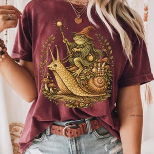 May include: Burgundy t-shirt featuring a whimsical illustration of a frog wizard riding a snail. The frog wears a pointed hat and cape, holding a staff. The snail has a basket of mushrooms. The design is surrounded by foliage.