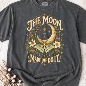 Op de afbeelding: Een antracietgrijs t-shirt met een grafische print van een halve maan en een nachtvlinder omringd door bloemen. De tekst THE MOON MADE ME DO IT staat in een crèmekleurig schreeflettertype boven en onder de illustratie.