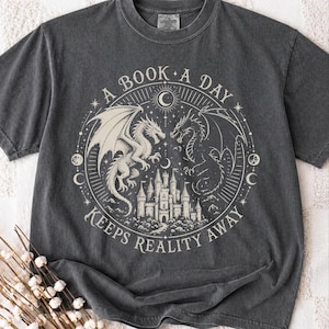 Puede incluir: Una camiseta de manga corta gris carbón con un gráfico circular de color crema. El diseño muestra dos dragones flanqueando un castillo con el texto A BOOK A DAY KEEPS REALITY AWAY dispuesto en un círculo alrededor de la ilustración central.