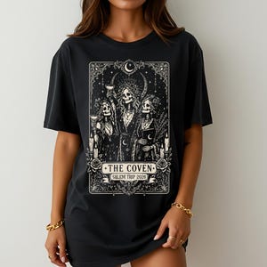 May include: A black t-shirt featuring a white graphic print of three skeletons in robes, holding glasses and a book. The design includes crescent moons, stars, candles, and roses, with the text THE COVEN and SALEM TRIP 2026 in a banner.