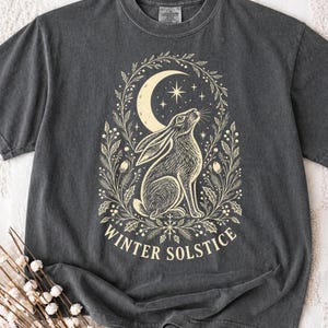 以下が含まれることがあります： チャコールグレーの半袖Tシャツ。三日月と星の下に座るウサギと、それを取り囲む植物のリースがクリーム色で描かれています。ウサギの下には「WINTER SOLSTICE」という文字がアーチ状にプリントされています。