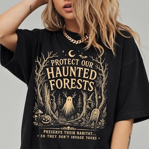 Op de afbeelding: Een zwart T-shirt met korte mouwen en een gouden grafische print van een bosscène met een geest, een pompoen en een grafsteen. De tekst luidt: PROTECT OUR HAUNTED FORESTS, PRESERVE THEIR HABITAT... SO THEY DON'T INVADE YOURS.